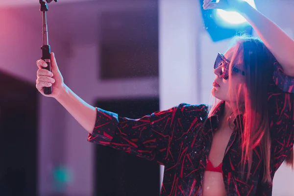 Atractiva mujer joven en camisa a cuadros tomando selfie usando monópodo con teléfono inteligente. — Foto de Stock