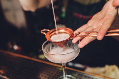 farklı bardaklar, metal zımbırtı ve ölçü kabı tezgahın üzerinde duruyor. Arka planda bulanık barmen.