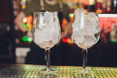 farklı bardaklar, metal zımbırtı ve ölçü kabı tezgahın üzerinde duruyor. Arka planda bulanık barmen.