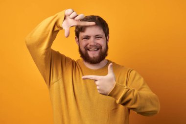 Neşeli, tıraşsız bir adam iki eliyle çerçeve işareti yapar, fotoğraflanmaya hazırlanır, günlük kırmızı tişört giyer, beyaz arka plana karşı durur. Hayata parlak bir açıdan bak..