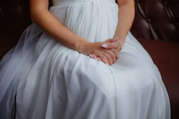 A girl in a white dress is sitting on a soft armchair, holding her hands in her lap. Hands of the bride, white dress, bride. Wedding morning. — Stock Photo, Image