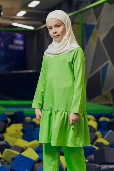 Seitenporträt eines kleinen muslimischen Mädchens, das einen Hijab trägt. Konzept muslimischer Kleidung für Kinder. vor dem Hintergrund des Spielplatzes. — Stockfoto