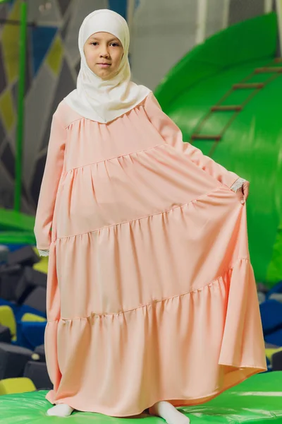 Seitenporträt eines kleinen muslimischen Mädchens, das einen Hijab trägt. Konzept muslimischer Kleidung für Kinder. vor dem Hintergrund des Spielplatzes. — Stockfoto