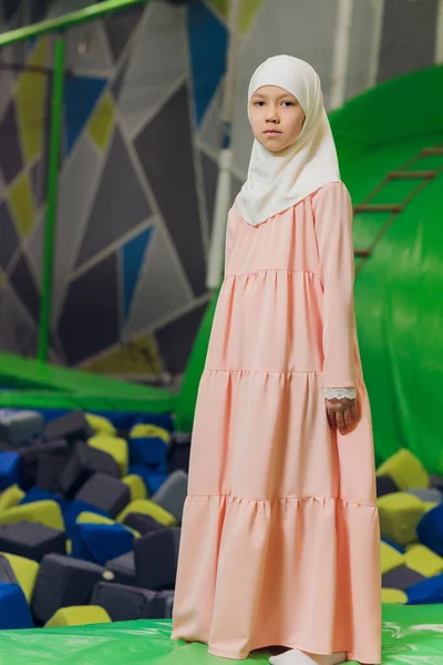 Seitenporträt eines kleinen muslimischen Mädchens, das einen Hijab trägt. Konzept muslimischer Kleidung für Kinder. vor dem Hintergrund des Spielplatzes. — Stockfoto