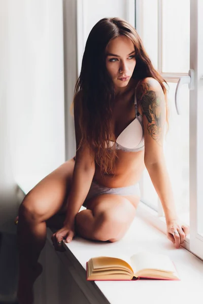 Mujer joven en ropa interior en la habitación. chica en lencería de pie cerca de la ventana. chica leyendo libro al lado de la ventana. —  Fotos de Stock