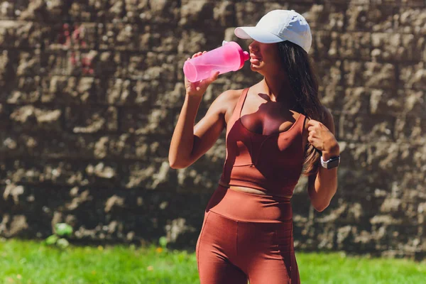 Γυμνή κοιλιά και λεπτό σχήμα αθλητικής γυναίκας, κρατώντας ένα μπουκάλι νερό και φορώντας ένα περικάρπιο. Πρωινή συνεδρία στην πόλη. Χύνεται πάνω της. — Φωτογραφία Αρχείου