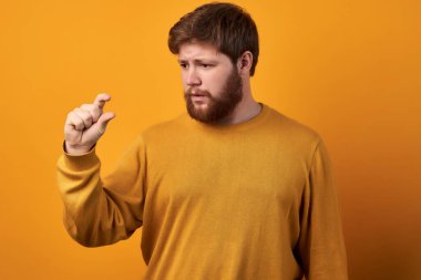 Şaşırmış sakallı erkeğin yatay pozu çok küçük bir şey gösteriyor. Beyaz arka planda izole edilmiş sıradan bir kot gömlek giyiyor. Şok olmuş modacı adam sefil maaşının büyüklüğünü gösteriyor..