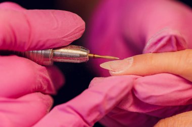 Bir kadın kuaförde manikür yapıyor, usta çalışırken tırnaklarını kesiyor, manikür yapıyor. Eldivenli eller.