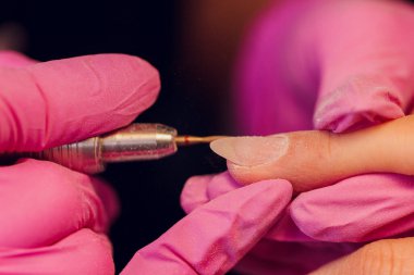 Bir kadın kuaförde manikür yapıyor, usta çalışırken tırnaklarını kesiyor, manikür yapıyor. Eldivenli eller.