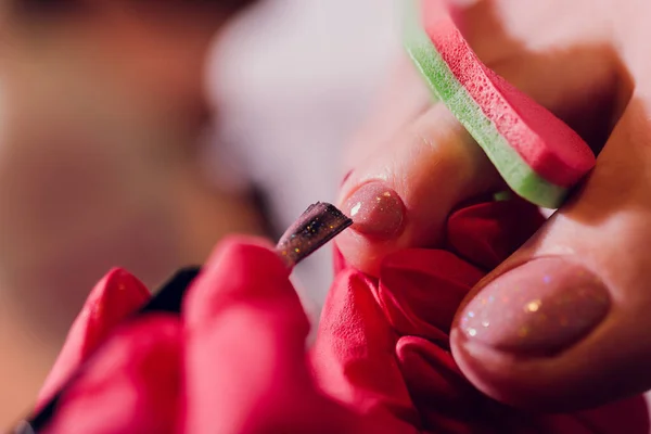 Le mani in guanti si preoccupano di unghie di piede delle donne. Pedicure, manicure concetto di salone di bellezza. Smalto per unghie di colore rosso. — Foto Stock