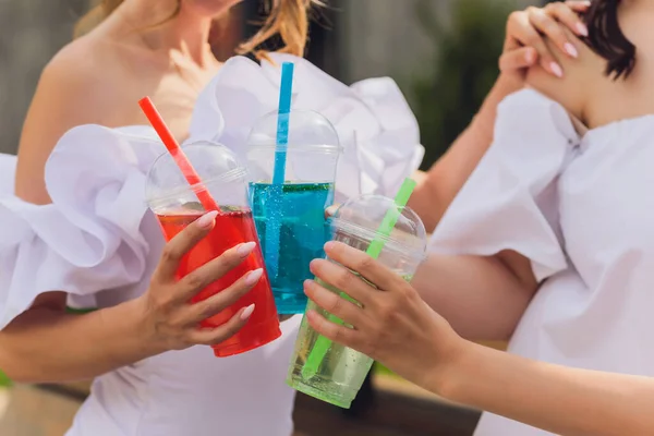 Three ladies cheering with tasty nectars with ice cubes, with black straws, palms decoration, in swim pool, transparent clean clear blue water, sun shines, smooth tanned skin, carefree festive mode. — Stock Photo, Image