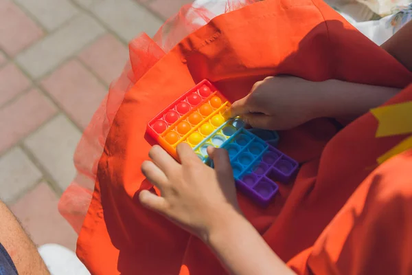 Niño anónimo sostiene en sus manos juguete sensorial pop y lo muestra delante de sí mismo.Arco iris color.Juguete antiestrés para niños y adultos. — Foto de Stock