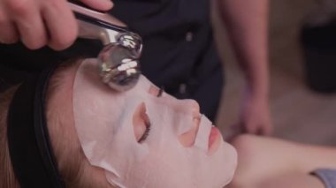 Çıplak omuzlu genç bir kadın, kahverengi arka planda tek başına dikilirken yüz masajı yapıyor ve başka tarafa bakıyor..