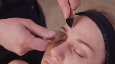 Çıplak omuzlu genç bir kadın, kahverengi arka planda tek başına dikilirken yüz masajı yapıyor ve başka tarafa bakıyor..