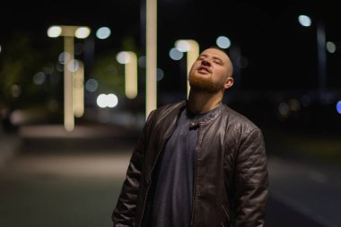 Bir adam sokak kenti boyunca yürür. Arka planda fenerlerle süslenmiş bir caddede genç bir adamın portresi var..