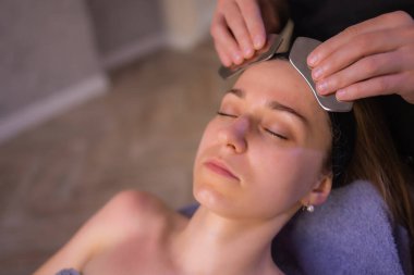 Çıplak omuzlu genç bir kadın, kahverengi arka planda tek başına dikilirken yüz masajı yapıyor ve başka tarafa bakıyor..