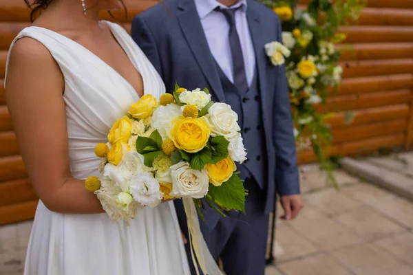 Novia y el novio de pie sobre hierba verde y la celebración de un ramo de flores blancas y amarillas con verde. —  Fotos de Stock