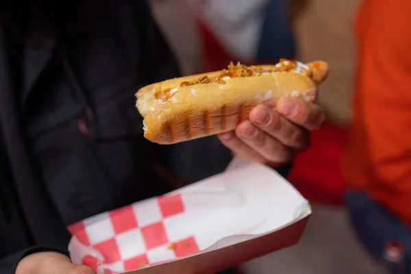 hand holds fried sausage in dough with ketchup in paper red white packaging on the street.