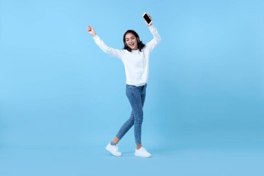 Mutlu gülümseyen Asyalı kadın kablosuz kulaklık takıyor müzik dinliyor ve mavi arka planda dans ediyor..