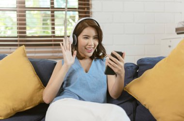 Mutlu Asyalı kadın akıllı telefon videosu kullanıyor. Oturma odasındaki kanepede sanal görüşme yapıyor..