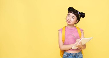 Genç Asyalı kız sarı arka planda izole edilmiş bir not defterine bir şeyler yazıp düşünüyor..