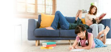 Relax Asian parent reading a book on sofa and daughter painting art in living room at home. panoramic background.	