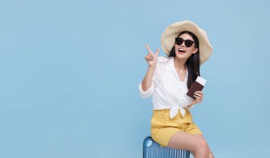 Happy smiling Asian woman dressed in summer clothes and wearing hat with luggage enjoying their summer vacation and pointing finger a copy space on bright blue background.	