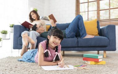 Rahatla Asyalı ebeveyn kanepede kitap okuyor ve kız evdeki oturma odasında resim yapıyor. panoramik arkaplan.