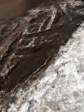 Şehir caddesinde koyu kahverengi tonlarda kirli eriyen kar ve çamur. Çamurlu kış karı, ayak izleri ve su birikintileri, kentsel mevsimlik arka plan. Soğuk hava, erime, kaygan yol koşulları ve kış tehlikeleri kavramı.