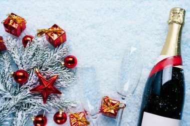 Noel ve Yeni Yıl kutlamaları sahnesinde bir şişe şampanya, iki boş şampanya bardağı, kırmızı bayram süsleri, buzlu çam dalları ve karlı arka planda düzenlenmiş küçük hediye kutuları.