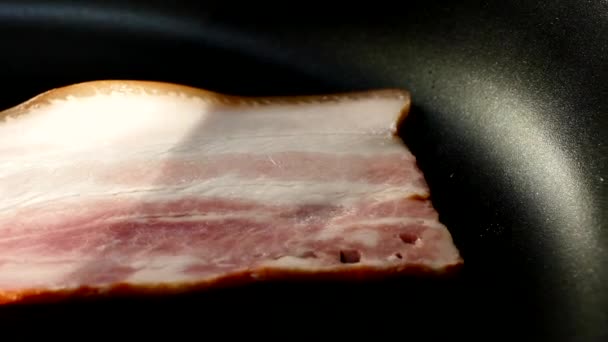 Freír el tocino en una sartén. Una rebanada de tocino de cerdo se prepara en su propia grasa. Capa de carne y manteca. Primer plano en la cocina. — Vídeos de Stock