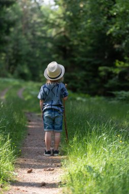 Elinde sopayla orman yolunda duran fötr şapkalı genç bir çocuk..