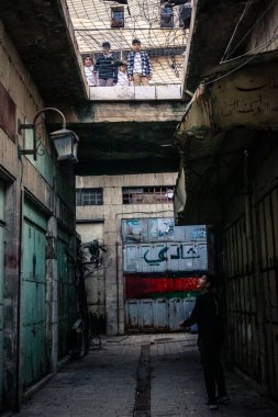 Hebron, Filistin Bölgesi, 31 Ocak 2026 Dikey Ayrımcılık. Üst katlardaki İsrailli göçmen çocuklar Filistin sokaklarına bakıyor. Metal ağ pazarı fırlatılan nesnelerden korur.