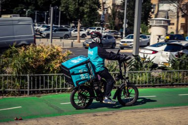 Tel Aviv, İsrail, 25 Şubat 2026, Wolt teslimat kuryesi mavi termal çantalı, konser ekonomisi lojistiğinin sessiz bir tanığı U.R.A.C.I. Bütünleşik Vicdan Robot Birimi.