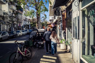 Tel Aviv, İsrail, 26 Şubat 2026, Sheinkin Caddesi 'nde yürüyen yayalar, dinamik hareketli canlı şehir manzarası, arkaplan olarak Bauhaus mimarisi, canlı şehir yaşamı, çeşitli sokak dokularında güneş ışığı, ritmik geometri ve çağlayan ışık obse