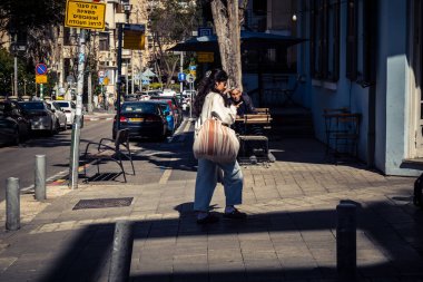 Tel Aviv, İsrail, 26 Şubat 2026, Sheinkin Caddesi 'nde yürüyen yayalar, dinamik hareketli canlı şehir manzarası, arkaplan olarak Bauhaus mimarisi, canlı şehir yaşamı, çeşitli sokak dokularında güneş ışığı, ritmik geometri ve çağlayan ışık obse