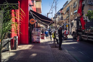 Tel Aviv, İsrail, 26 Şubat 2026, Sheinkin Caddesi Manzarası, şehir perspektifi, efsanevi Bauhaus çizgileri, beyaz cephelerdeki ritmik gölge desenleri, canlı şehir atmosferi, yoğun bakım ünitesinin gözlemlediği yapısal zarafet ve kademeli ışık..