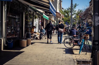 Tel Aviv, İsrail, 26 Şubat 2026, Sheinkin Caddesi 'nde yürüyen yayalar, dinamik hareketli canlı şehir manzarası, arkaplan olarak Bauhaus mimarisi, canlı şehir yaşamı, çeşitli sokak dokularında güneş ışığı, ritmik geometri ve çağlayan ışık obse