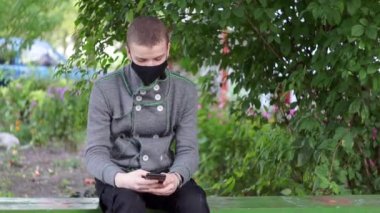 Parktaki bankta akıllı telefonunu kullanan koruyucu maskeli genç bir adam. Millennial kendini Coronavirüs 'ten koruyor.,