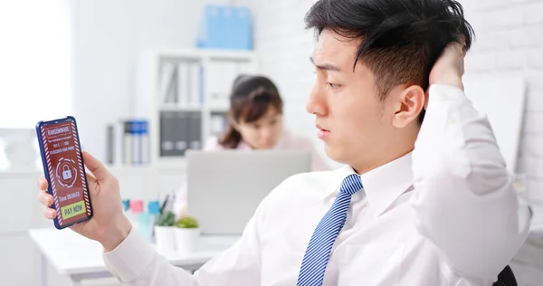 アジア人は若いビジネスマンがオフィスの画面上でランサムウェア攻撃の言葉でスマートフォンを見ているのを心配 — ストック写真