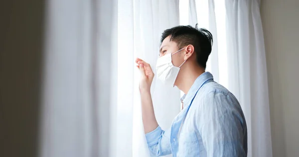 亚洲年轻人戴着口罩 因患了流行性感冒而呆在家里自我隔离 他朝窗外看了看 — 图库照片