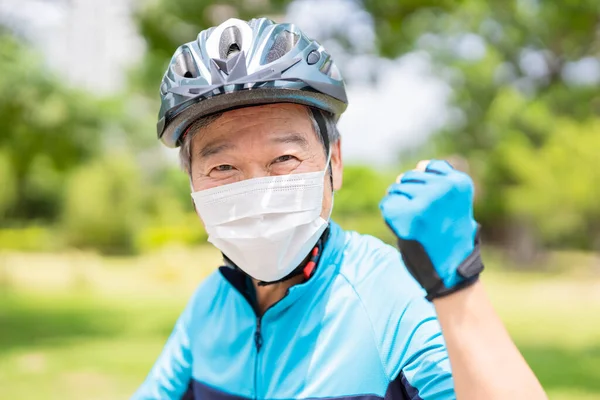 close up of smiling asian elder man wearing helmet and face mask with bicycle is looking to you - raise fist gesture