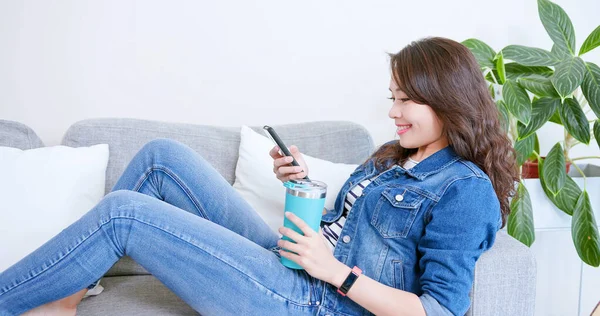 亚洲女人躺在沙发上 在家里用智能手机 用可重复使用的生态友好型生态瓶喝水 — 图库照片