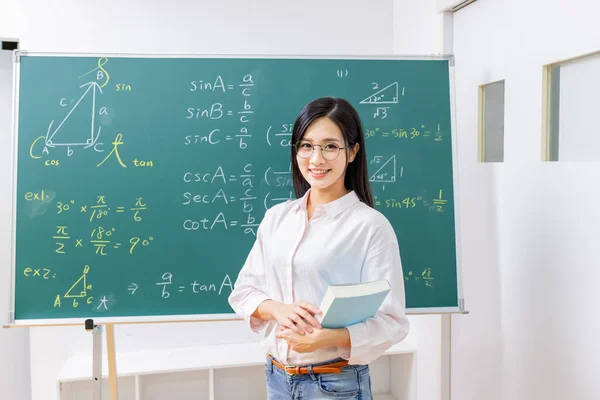 asian senior high school female math teacher hold books and smile at you in front of blackboard at classroom