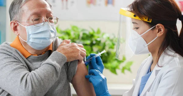 手袋と隔離マスクをしたアジア系女性医師が入院中の高齢男性患者の肩にワクチンを作っています — ストック写真