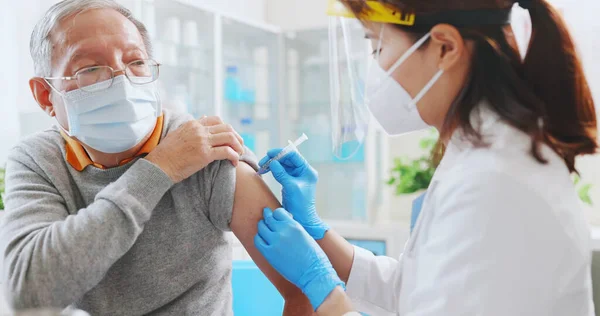 asian female doctor wearing gloves and isolation mask is making a vaccination in the shoulder of senior male patient at hospital