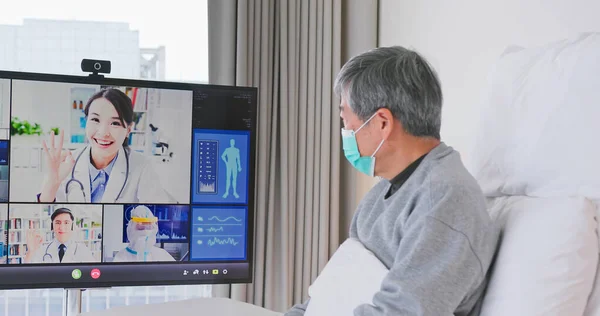 Telemedicine concept - asian senior man patient lying on bed has video call with doctor team by big screen TV in a hospital ward or at home