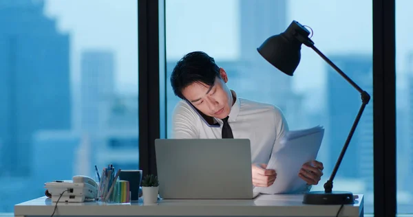 アジアの若いビジネスマンはオフィスで懸命に仕事をしながらノートパソコンを持ったスマートフォンで話す — ストック写真