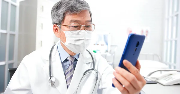 远程医疗概念 亚洲资深男医生在诊所通过带口罩的手机视频聊天向病人解释这种疾病 — 图库照片