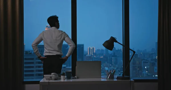 心配してるアジアの若いビジネスマンは夜勤の残業中窓の外を見てる — ストック写真